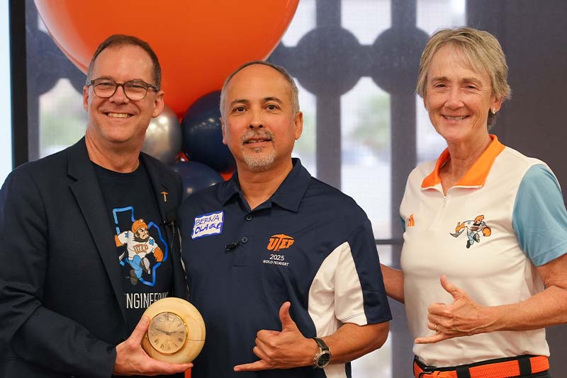 Three attendees pose together during a UTEP Engineering homecoming recognition event, with one holding a commemorative award while standing in front of orange, blue, and silver balloon decorations and making hand gesture signs for the camera.